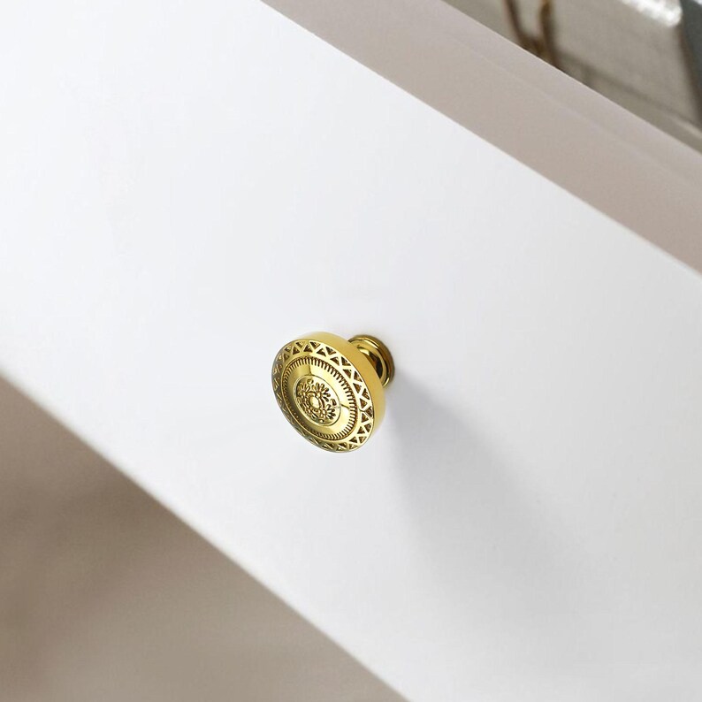 May include: A gold-colored cabinet knob with an ornate, circular design. The knob is mounted on a white surface, and the design features a central floral motif surrounded by geometric patterns. The hardware is likely for furniture or cabinetry.