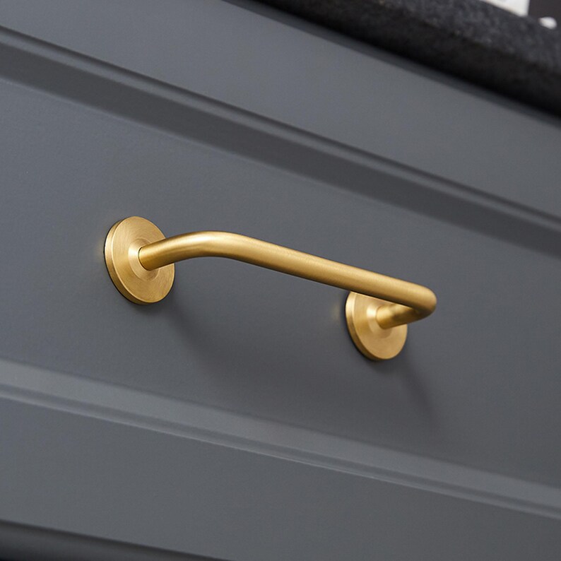 May include: A gold-colored, curved drawer pull with circular mounting plates on a gray cabinet door. The handle is a simple, elegant design, adding a touch of sophistication to the furniture. The hardware is made of metal.
