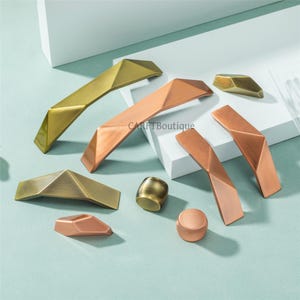 May include: A collection of geometric cabinet hardware in various metallic finishes, including gold, copper, and bronze. The hardware features angular designs and includes handles and knobs. The background is a light blue surface with white geometric shapes.