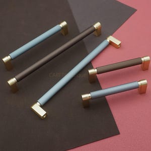 May include: A collection of cabinet pulls in various colors. The pulls have a ribbed center section in shades of blue and brown, with gold-toned rectangular end pieces. The pulls are arranged on a brown and pink background.