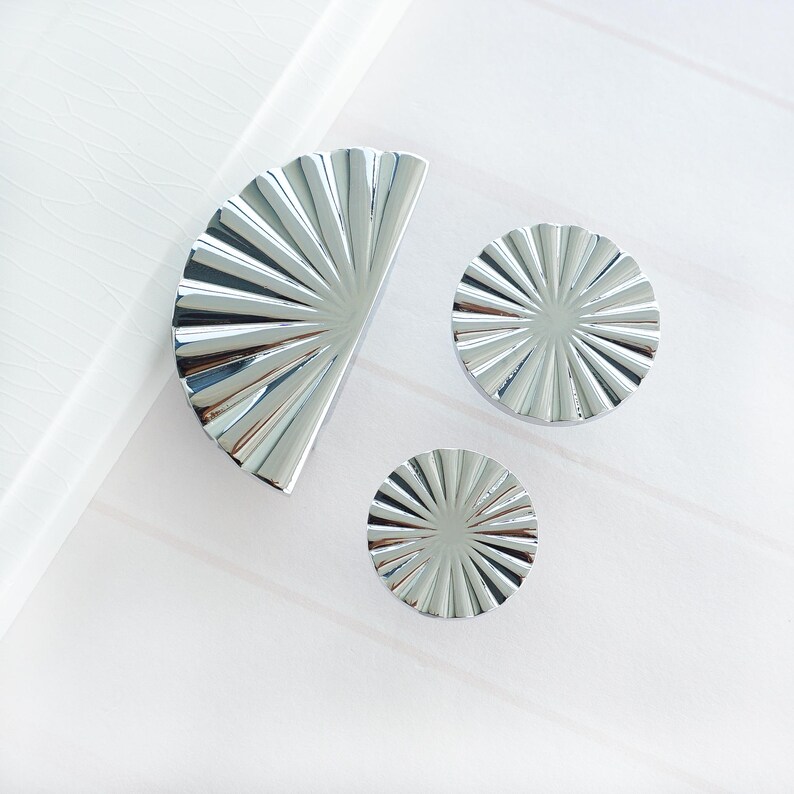 May include: Three silver cabinet knobs in a fan-like design. One is semi-circular, and the others are circular. The knobs have a ridged pattern radiating from the centre. The shiny metal knobs would suit a modern kitchen or furniture.