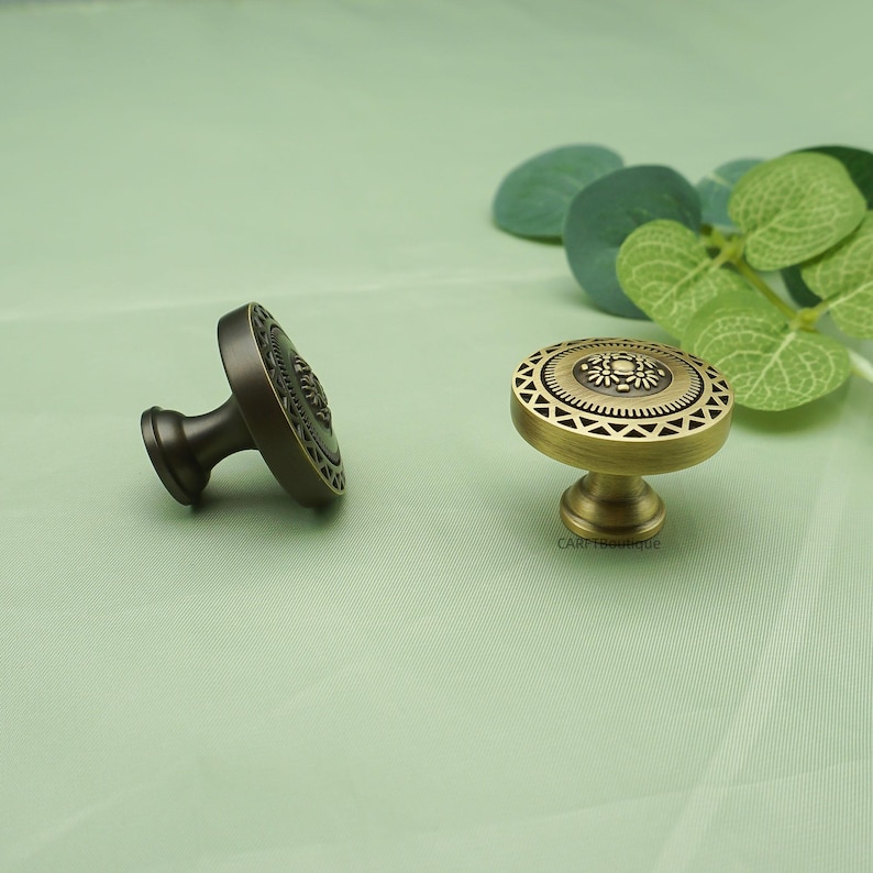 May include: Two decorative cabinet knobs. One is bronze-colored, the other is gold-colored. Both knobs have a circular design with intricate patterns. The knobs are likely for furniture or home decor.