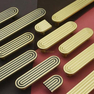 以下が含まれることがあります： 金色のキャビネットの取っ手とノブの詰め合わせ。取っ手は長方形で、一連の平行な溝と黒いアクセントがあります。ノブは同様のデザインの半円形です。ハードウェアは茶色と赤色の表面に配置されています。