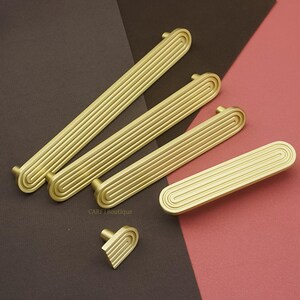 以下が含まれることがあります： 金色のキャビネットプルとノブのコレクション。プルは溝付きの長方形のデザインで、端は丸みを帯びています。ノブは同様の溝付きパターンを持つハート型です。茶色とピンクの背景に配置されています。