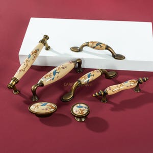 May include: A selection of antique-style cabinet handles and knobs. The hardware has a cream ceramic centre with a blue bird and floral design, set on a bronze-toned metal base. The handles are in various sizes and shapes, suitable for cabinets.