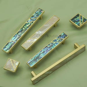 May include: A collection of cabinet handles and knobs in gold with abalone shell and mother-of-pearl inlays. The hardware is displayed on a green surface. The handles and knobs are for cabinets.