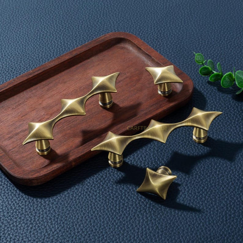 以下が含まれることがあります： 星型のノブと湾曲したハンドルを含む、アンティーク真鍮のキャビネットハードウェアのコレクション。 ハードウェアは、濃い青色の革のような表面の木製トレイに表示されます。 ハンドルはユニークで角張ったデザインです。