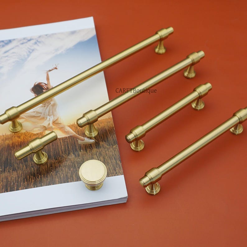 Pode incluir: Uma cole&ccedil;&atilde;o de ferragens de arm&aacute;rio de cor lat&atilde;o, incluindo v&aacute;rias puxadores de barra, um bot&atilde;o e um puxador menor. O hardware &eacute; exibido em uma revista branca e uma superf&iacute;cie de cor ferrugem.