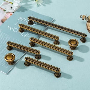 Może przedstawiać: Asortyment antycznych mosiężnych okuć do szafek, w tym kilka długich uchwytów i okrągłych gałek. Okucia charakteryzują się szczegółowym, żebrowanym wzorem i ciepłym, postarzanym wykończeniem. Przedmioty są ułożone na jasnoniebieskiej powierzchni.