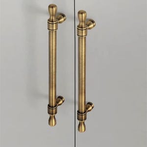 K&ouml;nnte beinhalten: Zwei antike Messing-Schrankgriffe. Jeder Griff hat einen langen, zylindrischen K&ouml;rper mit dekorativen Zierkn&ouml;pfen oben und unten. Sie sind auf einer hellgrauen Oberfl&auml;che montiert, was auf eine Schrankt&uuml;r hindeutet.