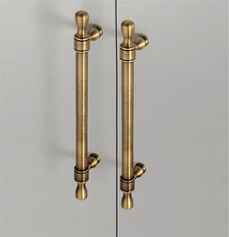 K&ouml;nnte beinhalten: Zwei antike Messing-Schrankgriffe. Jeder Griff hat einen langen, zylindrischen K&ouml;rper mit dekorativen Zierkn&ouml;pfen oben und unten. Sie sind auf einer hellgrauen Oberfl&auml;che montiert, was auf eine Schrankt&uuml;r hindeutet.