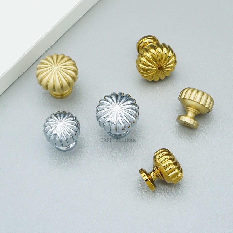 May include: A collection of decorative cabinet knobs in gold and silver finishes. The knobs feature a fluted, floral-like design. The hardware is displayed on a light gray surface. The knobs are suitable for furniture and home decor projects.