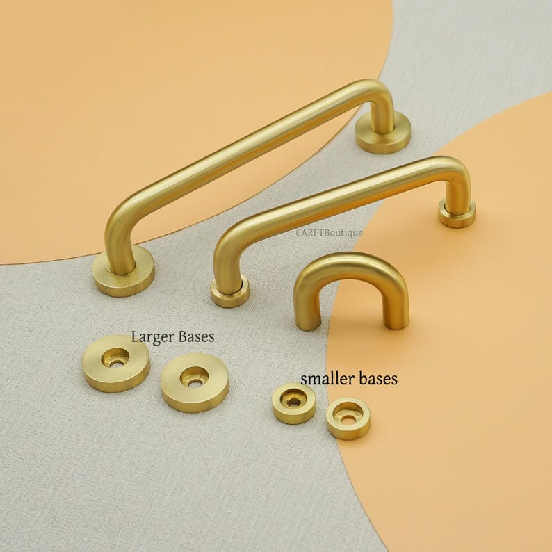 May include: A selection of brass-toned cabinet handles and base plates. The handles come in different sizes and designs, including a straight bar and a curved shape. The image also shows the text "Larger Bases" and "smaller bases".