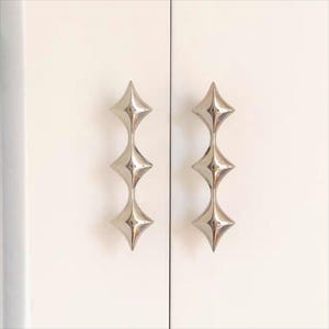 以下が含まれることがあります： スターバーストデザインの2つのシルバーキャビネットプル。各プルは、細く湾曲したステムで接続された3つのダイヤモンド型の要素を備えています。プルは白いキャビネットドアに取り付けられています。