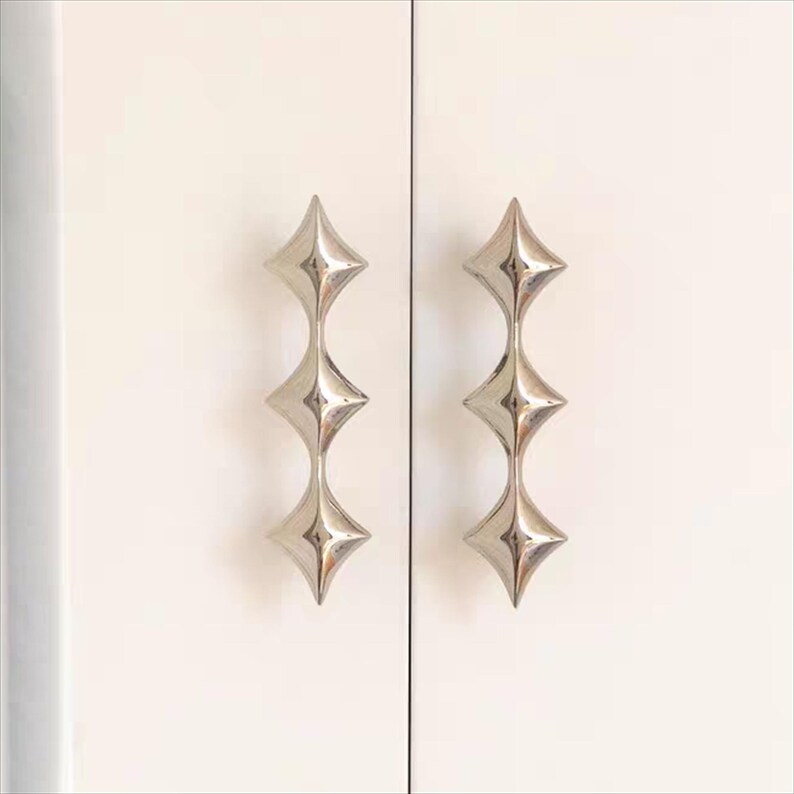 以下が含まれることがあります： スターバーストデザインの2つのシルバーキャビネットプル。各プルは、細く湾曲したステムで接続された3つのダイヤモンド型の要素を備えています。プルは白いキャビネットドアに取り付けられています。