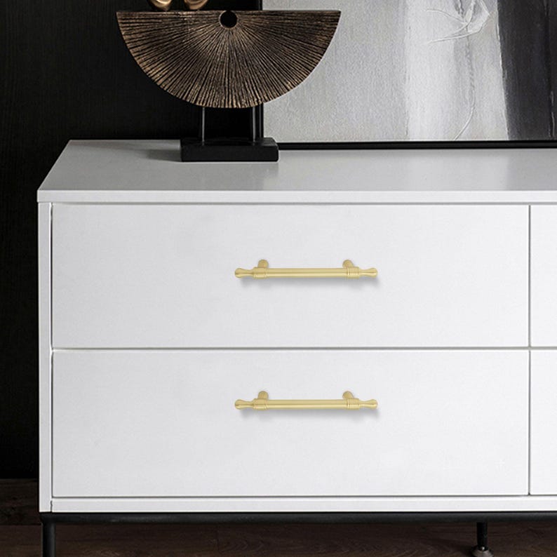 K&ouml;nnte beinhalten: Eine wei&szlig;e Kommode mit zwei Schubladen, jede mit einem goldfarbenen Stangengriff. Die Griffe sind horizontal ausgerichtet. Auf der Kommode befindet sich eine dekorative Bronzeskulptur.