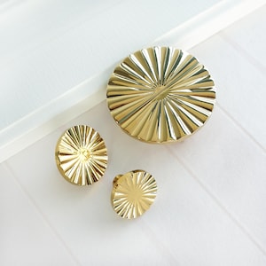 May include: Three gold-coloured cabinet knobs and a larger matching round piece. The knobs and round piece have a sunburst design with radial grooves. The items are on a white surface.