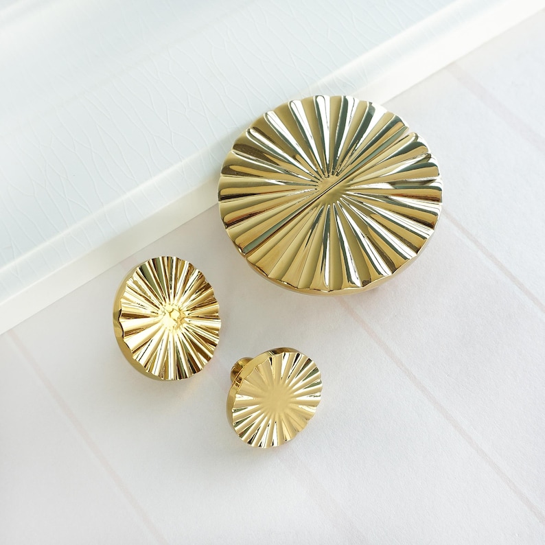 May include: Three gold-coloured cabinet knobs and a larger matching round piece. The knobs and round piece have a sunburst design with radial grooves. The items are on a white surface.