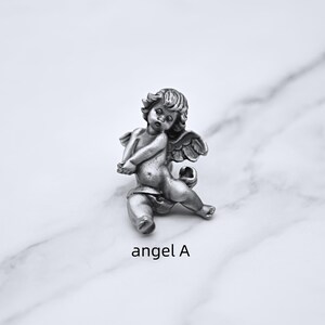 Peut inclure: Une figurine de ch&eacute;rubin argent&eacute; en position assise, avec des ailes d&eacute;taill&eacute;es et des cheveux boucl&eacute;s. Le ch&eacute;rubin repose sur une surface en marbre blanc. Le texte "angel A" est en dessous.