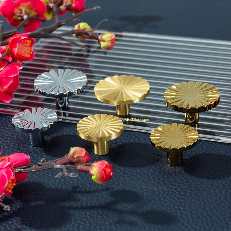 May include: A selection of decorative cabinet knobs in silver and gold. The knobs feature a round, fluted design, resembling a flower. They are displayed on a dark surface, with a floral arrangement in the background.