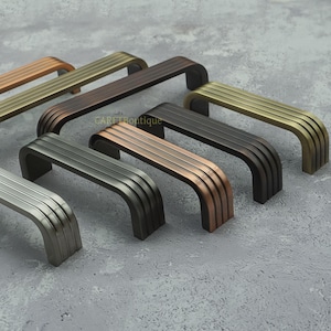 May include: A selection of cabinet handles in diverse metallic finishes, such as bronze, brass, and silver. Each handle has a curved design with parallel grooves, providing a modern look. The handles are displayed on a grey surface.