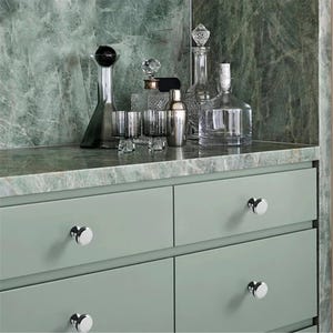 Peut inclure: Une collection de verrerie et de barware sur un comptoir en marbre. Comprend des carafes, des verres &agrave; cocktail et un shaker. Le meuble en dessous est peint en vert clair avec des boutons argent&eacute;s. Le fond est un mur en marbre vert.