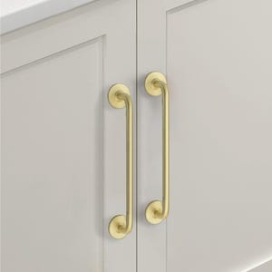 May include: Two gold-colored cabinet pulls with a curved design. The pulls are mounted on a light gray cabinet door. The pulls have a circular base and a curved handle, adding a touch of elegance to the cabinet.