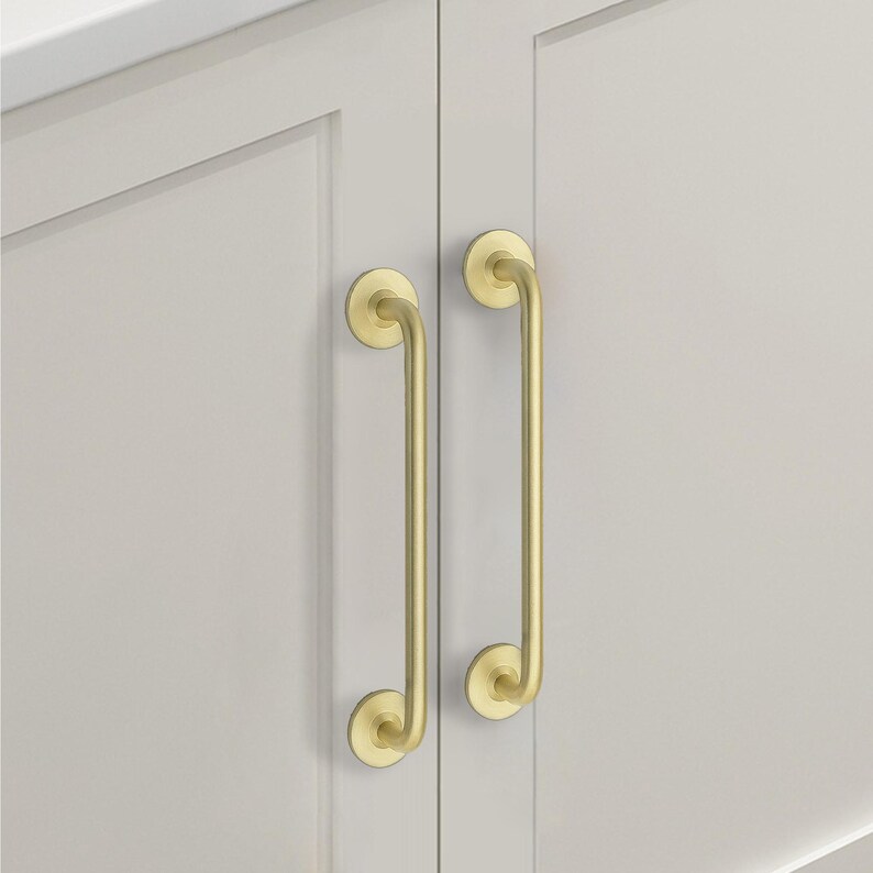 May include: Two gold-colored cabinet pulls with a curved design. The pulls are mounted on a light gray cabinet door. The pulls have a circular base and a curved handle, adding a touch of elegance to the cabinet.