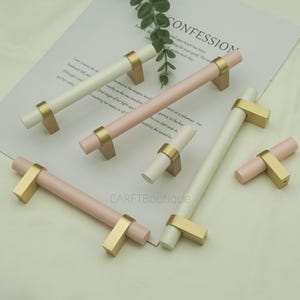 May include: A collection of cabinet pulls in various sizes and colors. The pulls have a cylindrical design with a gold-colored base and are available in white and pink. The word "CONFESSION" is visible in the background.