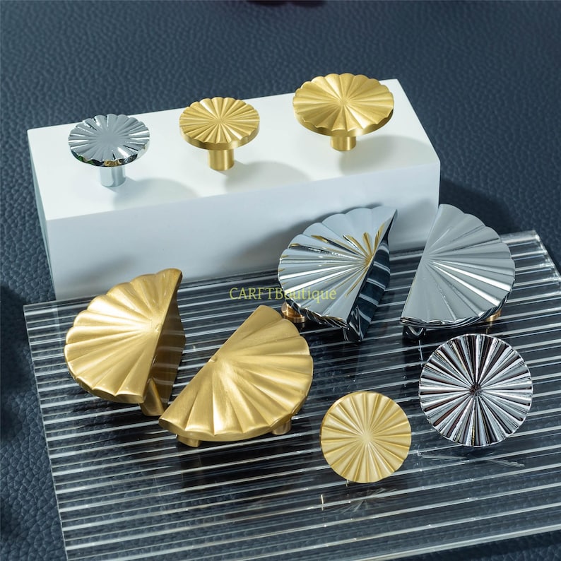 May include: A selection of decorative cabinet hardware, including gold and silver knobs and half-moon pulls. The knobs have a fluted, sunburst design. The pulls are half-circle shaped with a similar fluted pattern. The hardware is displayed on a white block and a clear, striped surface.
