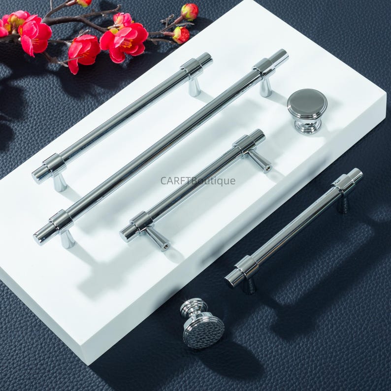 Peut inclure: Ensemble de quincaillerie de meubles chrom&eacute;e polie, comprenant des poign&eacute;es et des boutons. Les poign&eacute;es varient en longueur, et les boutons ont un design arrondi. Le mat&eacute;riel est pr&eacute;sent&eacute; sur une surface blanche avec un fond sombre, et une branche de fleurs rouges.
