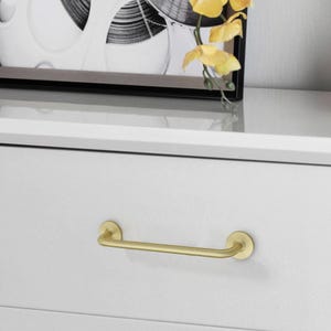 May include: A gold-colored drawer pull with a bar handle. The handle is mounted on a white drawer. The drawer pull has a simple, modern design. The drawer pull is made of metal.