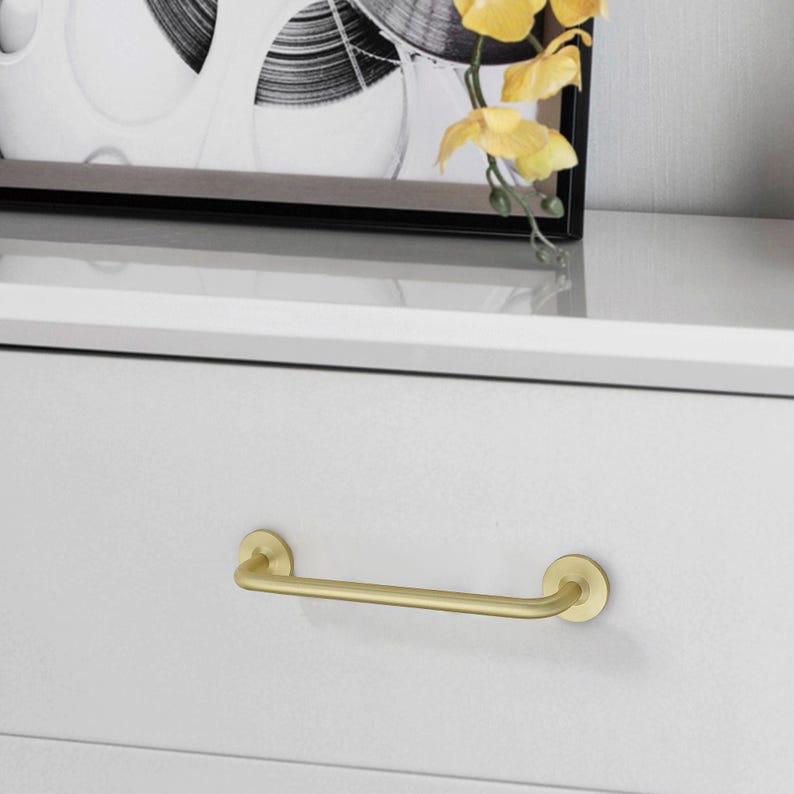 May include: A gold-colored drawer pull with a bar handle. The handle is mounted on a white drawer. The drawer pull has a simple, modern design. The drawer pull is made of metal.