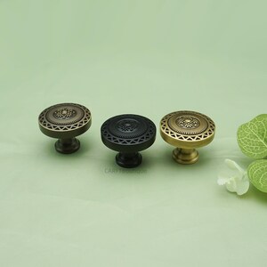 May include: Three decorative cabinet knobs in antique brass, black, and gold. Each knob features a circular design with a floral center and geometric border. The knobs are displayed on a light green background.