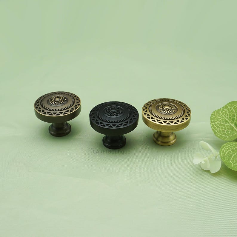 May include: Three decorative cabinet knobs in antique brass, black, and gold. Each knob features a circular design with a floral center and geometric border. The knobs are displayed on a light green background.