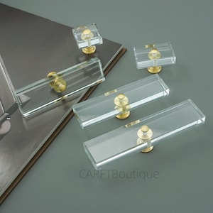 May include: Clear rectangular and square cabinet knobs with gold-toned bases. The knobs are made of a transparent material, reflecting light. The hardware is displayed on a grey surface. The text "CARFTBoutique" is visible.