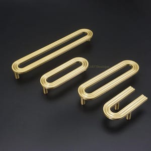 May include: Four gold-toned cabinet pulls in varying sizes, arranged on a dark background. The pulls have an oval design with a grooved texture, and are mounted on short, cylindrical posts. The overall aesthetic is modern and minimalist.