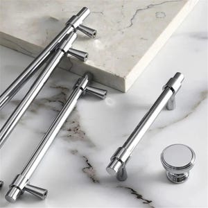 Peut inclure: Ensemble de quincaillerie de meubles en chrome poli, comprenant plusieurs poign&eacute;es de barre et un bouton rond. Les articles sont pr&eacute;sent&eacute;s sur une surface en marbre blanc et gris. Les poign&eacute;es ont un design cylindrique avec une poign&eacute;e en forme de T.