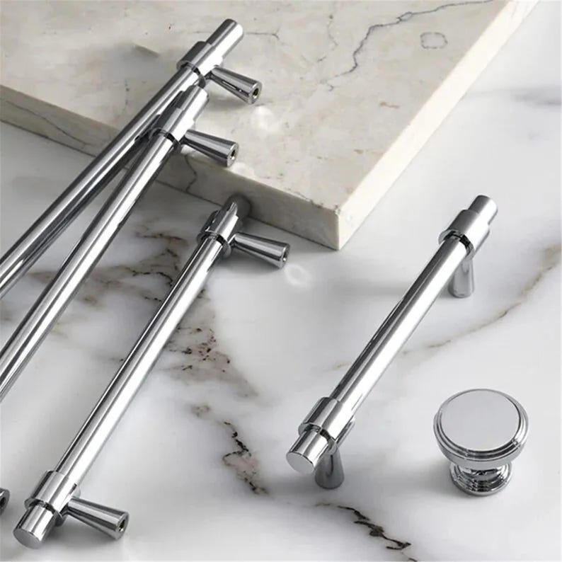 Peut inclure: Ensemble de quincaillerie de meubles en chrome poli, comprenant plusieurs poign&eacute;es de barre et un bouton rond. Les articles sont pr&eacute;sent&eacute;s sur une surface en marbre blanc et gris. Les poign&eacute;es ont un design cylindrique avec une poign&eacute;e en forme de T.