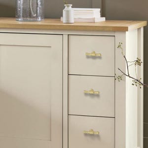 K&ouml;nnte beinhalten: Cremefarbener Schrank mit drei Schubladen, jede mit einem goldfarbenen Griff im Bambus-Stil. Der Schrank hat eine helle Holzplatte und befindet sich neben einer wei&szlig;en Platte. Eine Vase und Dekorationsgegenst&auml;nde befinden sich auf der Arbeitsplatte.