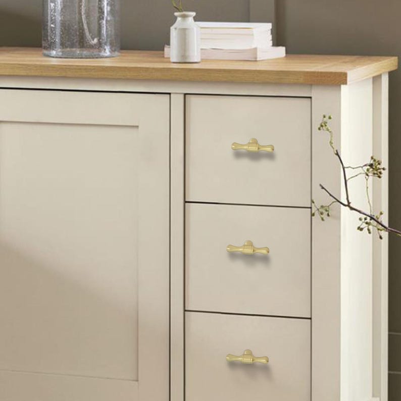 K&ouml;nnte beinhalten: Cremefarbener Schrank mit drei Schubladen, jede mit einem goldfarbenen Griff im Bambus-Stil. Der Schrank hat eine helle Holzplatte und befindet sich neben einer wei&szlig;en Platte. Eine Vase und Dekorationsgegenst&auml;nde befinden sich auf der Arbeitsplatte.