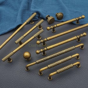 May include: An assortment of antique brass cabinet hardware, including handles and knobs. The handles vary in design, with ribbed, hammered, and smooth textures. The hardware is arranged on a dark surface with a blue cloth in the background.