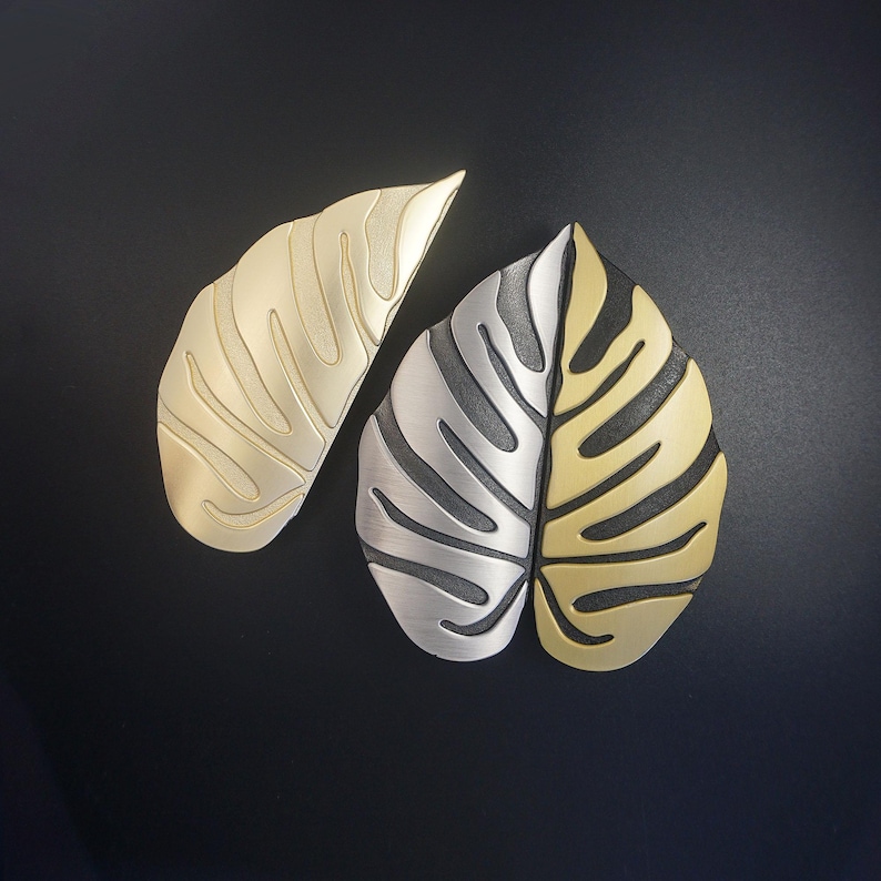 May include: Two metal leaf-shaped decorative objects. One is a solid gold color, the other is split, with one half silver and the other gold. The leaves have a textured, veined design and are set against a black background.
