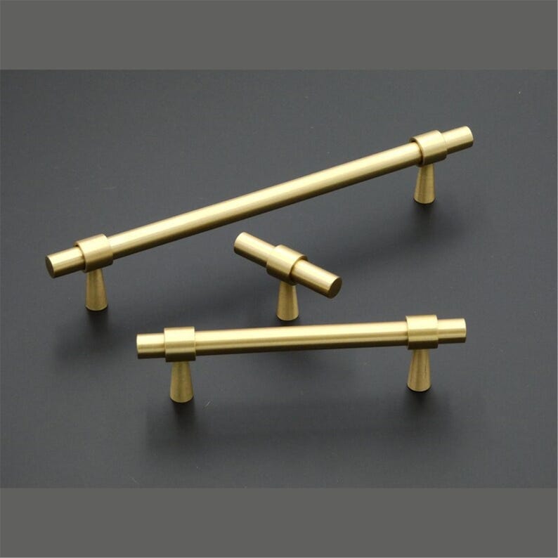 Peut inclure: Un ensemble de quatre poign&eacute;es de meubles de couleur laiton, de diff&eacute;rentes tailles. Les poign&eacute;es pr&eacute;sentent un design de barre cylindrique avec des pieds coniques. Le mat&eacute;riel est pr&eacute;sent&eacute; sur un fond sombre, soulignant la finition m&eacute;tallique.