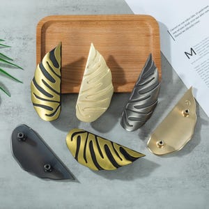 May include: A collection of decorative leaf-shaped cabinet pulls in various metallic finishes, including gold, brass, and gunmetal. The pulls are arranged on a wooden tray and a gray surface, showcasing their intricate design and potential for home decor.