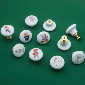 Könnte beinhalten: Eine Sammlung weißer Keramik-Schrankknöpfe mit verschiedenen Designs, darunter Teddybären, ein Einhorn und das Wort "love". Die Knöpfe haben Messing- oder goldfarbene Beschläge und sind auf grünem Grund angeordnet.