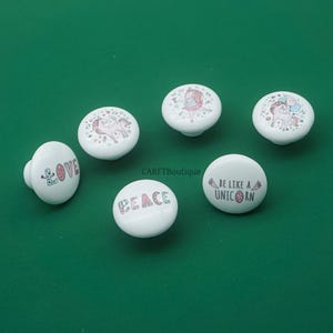 May include: White ceramic cabinet knobs with various designs, including unicorns, the word "LOVE", and the word "PEACE". The knobs have a round shape and are set against a green background. The text "BE LIKE A UNICORN" is also featured.