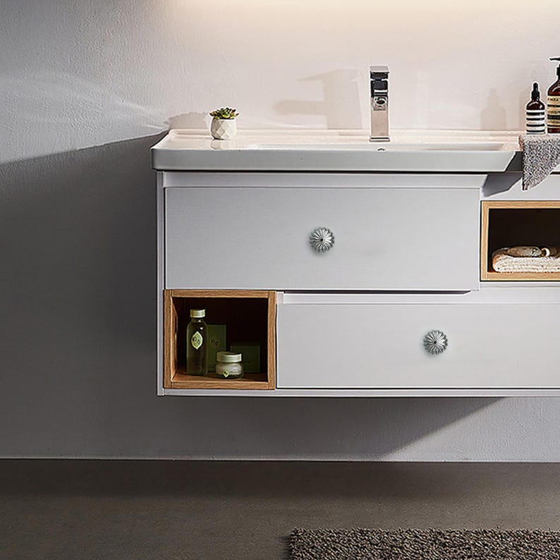 May include: A modern, wall-mounted bathroom vanity with a light gray finish. It features two drawers with decorative silver knobs, open wooden shelves, and a white countertop with a silver faucet. A small potted plant sits on the countertop.