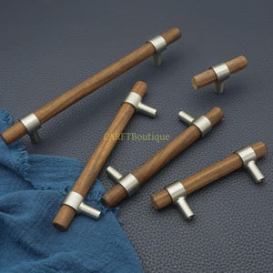 Puede incluir: Un conjunto de tiradores de madera para armarios con acabado de níquel cepillado. Los tiradores tienen un mango cilíndrico de madera y un anillo metálico que conecta con los postes de montaje. El texto "CARETBoutique" es visible.