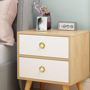 May include: A two-drawer bedside table with a light wood frame and white drawer fronts. The drawers have gold-coloured, sunburst-shaped knobs. A small digital clock, a framed photo, and a vase of flowers sit on top. The table has four angled legs.